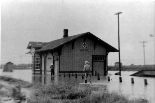 Man wading toward the small train depot building, which is surrounded by water (tracks not visible)