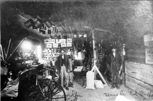 Dark interior of a hardware store, two men standing on either side at the back; a variety of hardware goods piled to the left