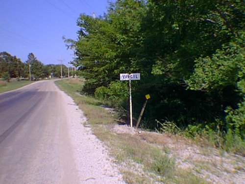 Strip of asphalt road to leading into Virgil, with a small sign bearing the town's name on the right