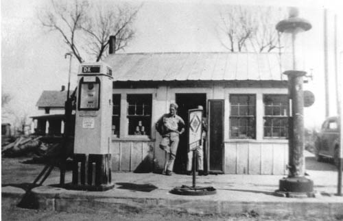 Two men lounge in front of a very small one-room buiding, with an older gas pump in the foreground