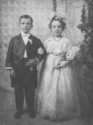 The boy and girl are very formally dressed, and posed as if for a traditional wedding photograph.